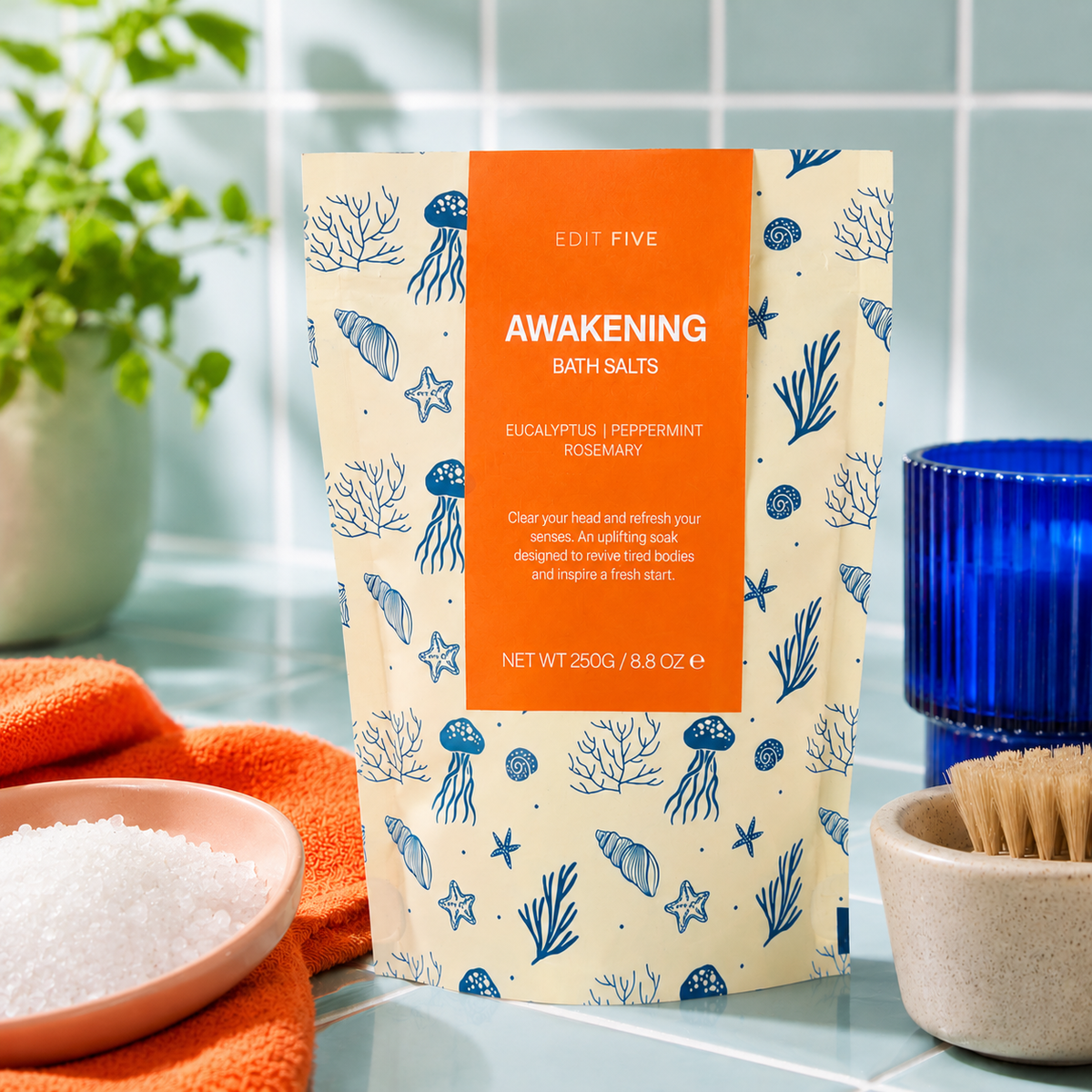 A stand-up pouch of Edit Five Awakening Bath Salts, featuring a cream background with blue sea-creature illustrations and a prominent orange label, set on a bathroom counter with towels, a blue cup, and a brush. Edit Five Awakening revitalising bath salts in sea-themed kraft packaging, designed to soothe tired muscles, refresh skin and create an invigorating minty-herbal bathing experience.