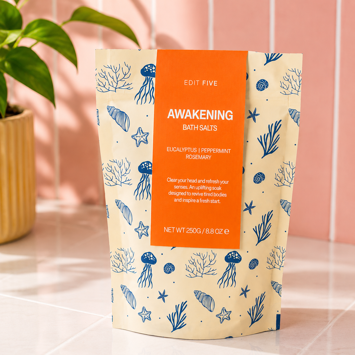 A stand-up pouch of Edit Five Awakening Bath Salts, featuring a cream-colored background with blue sea-themed illustrations and a prominent orange label, set on a tiled surface with a pink wall and a green plant in the background. Awakening bath salts with Epsom and Dead Sea salts, scented with eucalyptus, peppermint and rosemary essential oils for an energising, refreshing and uplifting bath.