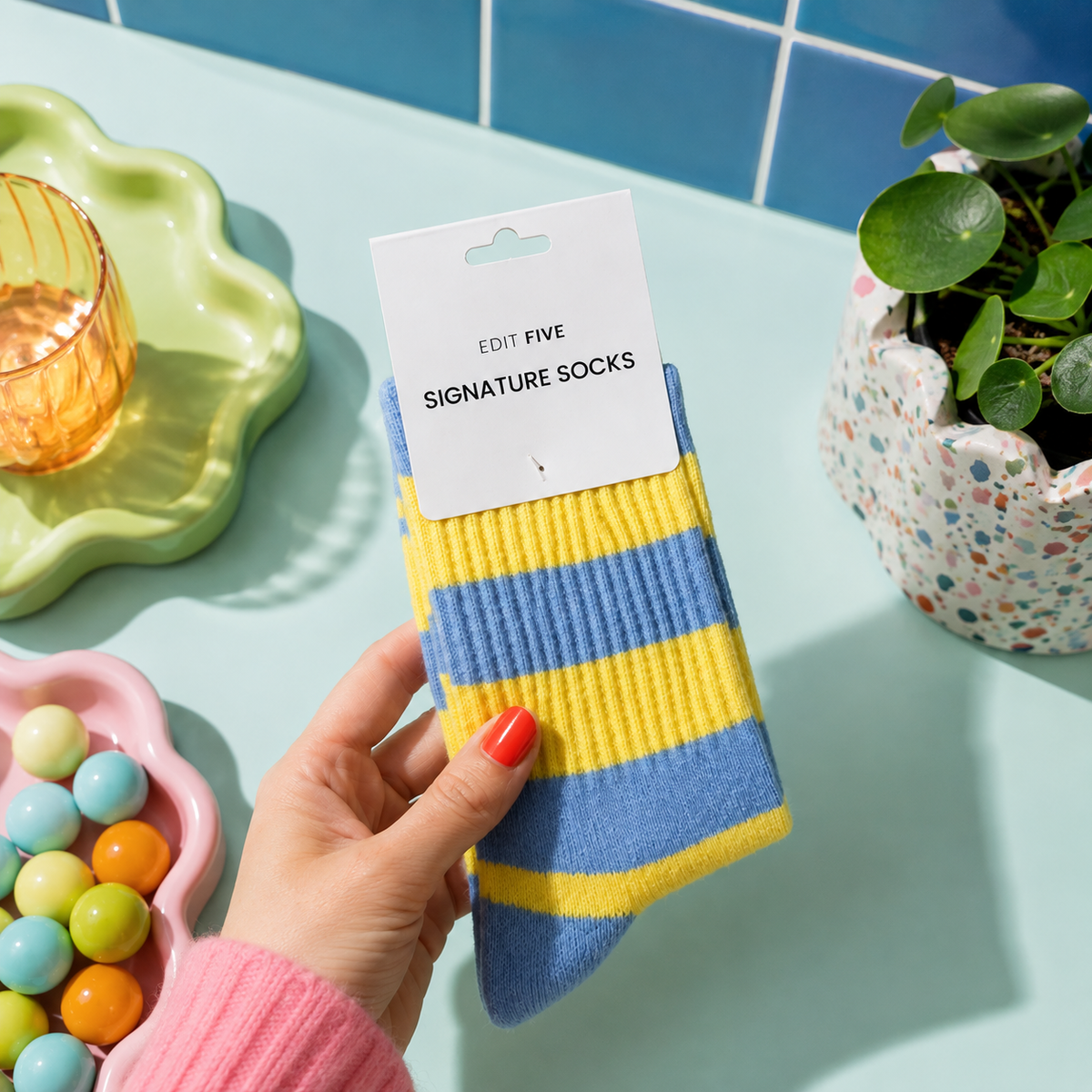A hand holds a pair of blue and yellow striped socks on a white paper hanger card, set against a styled tabletop with a plant, colorful candies, and a glass on a tray. Women’s blue and yellow striped socks made from a soft, warm and breathable wool-blend fabric, with a comfortable stretch fit for cosy everyday wear.