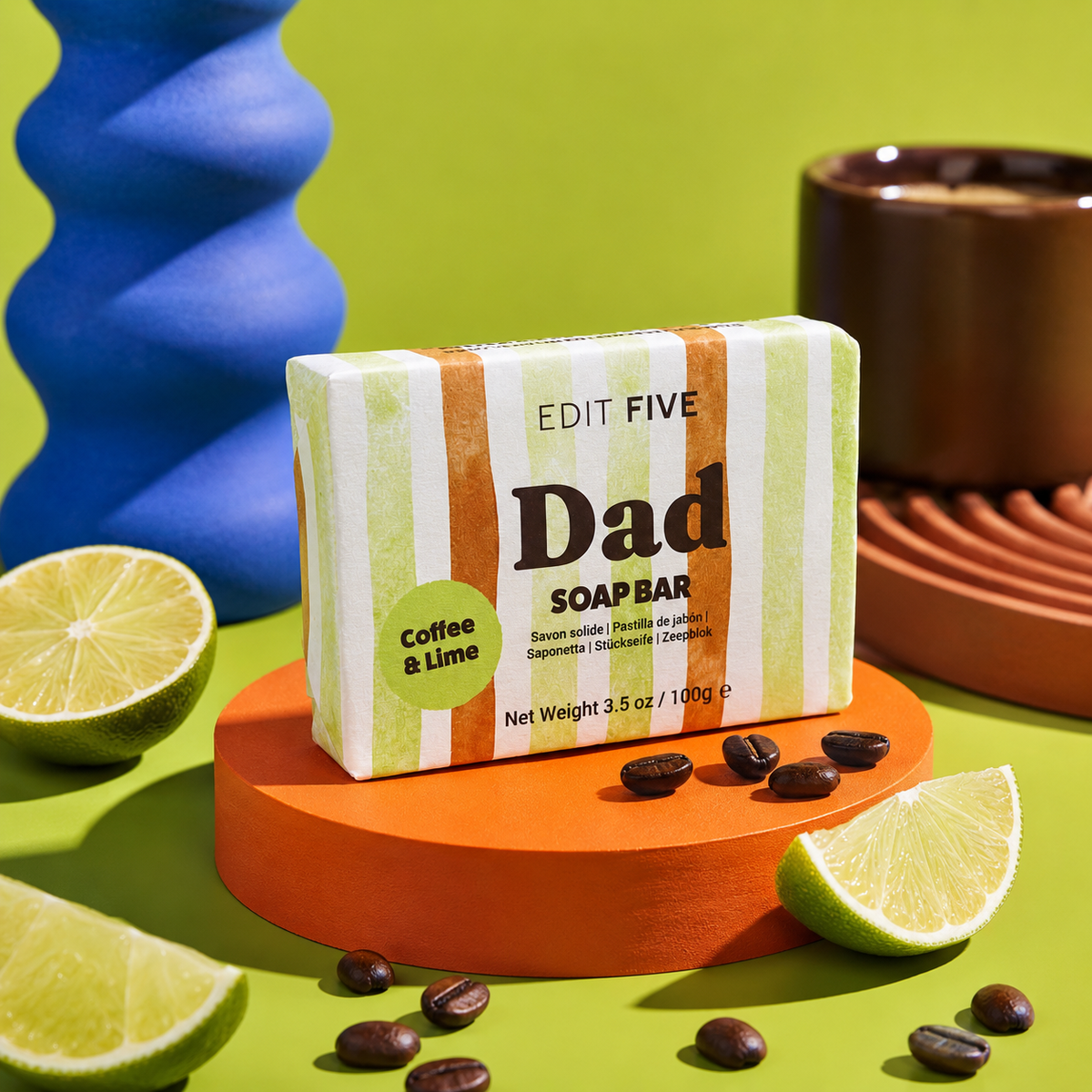 A bar of soap from Edit Five labeled 'Dad' and 'Coffee & Lime' is displayed on an orange pedestal. The soap bar has green, white, and orange vertical stripes. Luxury handmade men’s soap with exfoliating coffee and uplifting lime essential oil, creating a refreshing lather for everyday bathing, showering, skincare and grooming.