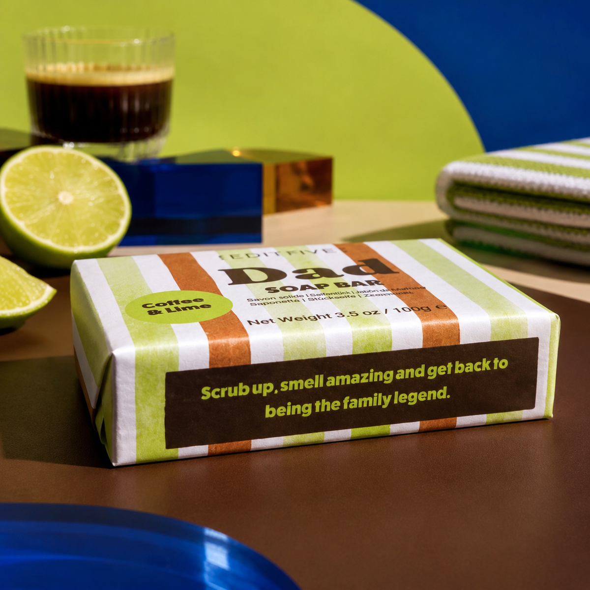 A boxed soap bar labeled 'Dad SOAP BAR' with green, white, and orange stripes sits on a tabletop. The package has a dark brown label reading 'Coffee & Lime' and a slogan. Edit Five coffee and lime soap bar designed especially for dads, featuring 100% natural essential oils and colourful packaging—a practical Father’s Day gift or birthday present.