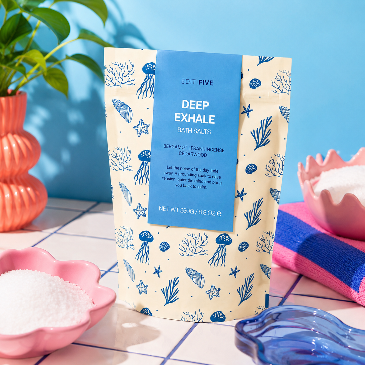 A stand-up pouch of Edit Five Deep Exhale Bath Salts stands on a tiled surface with pink bowls of white salt, a blue towel, and a plant in the background. Gift-ready Deep Exhale aromatherapy bath salts—a cruelty-free self-care gift, relaxation present, pamper-hamper addition or thoughtful wellbeing gift for bath lovers.
