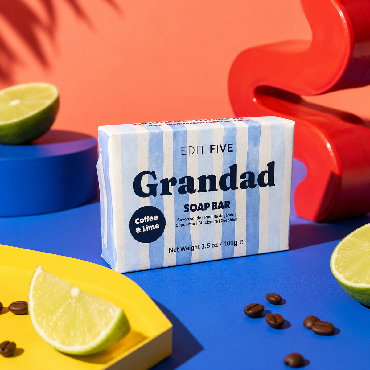 A boxed soap bar labeled 'Grandad SOAP BAR' with 'Coffee & Lime' sits on a bright blue surface, surrounded by lime wedges, coffee beans, and bold decorative props in orange, red, and yellow. The soap box is the main product, and no person or real environment is present, so this is Product/Packaging rather than Lifestyle. Luxury handmade soap for Grandad with exfoliating coffee and uplifting lime essential oil, creating a fresh, invigorating lather for everyday bathing, showering and men’s self-care.