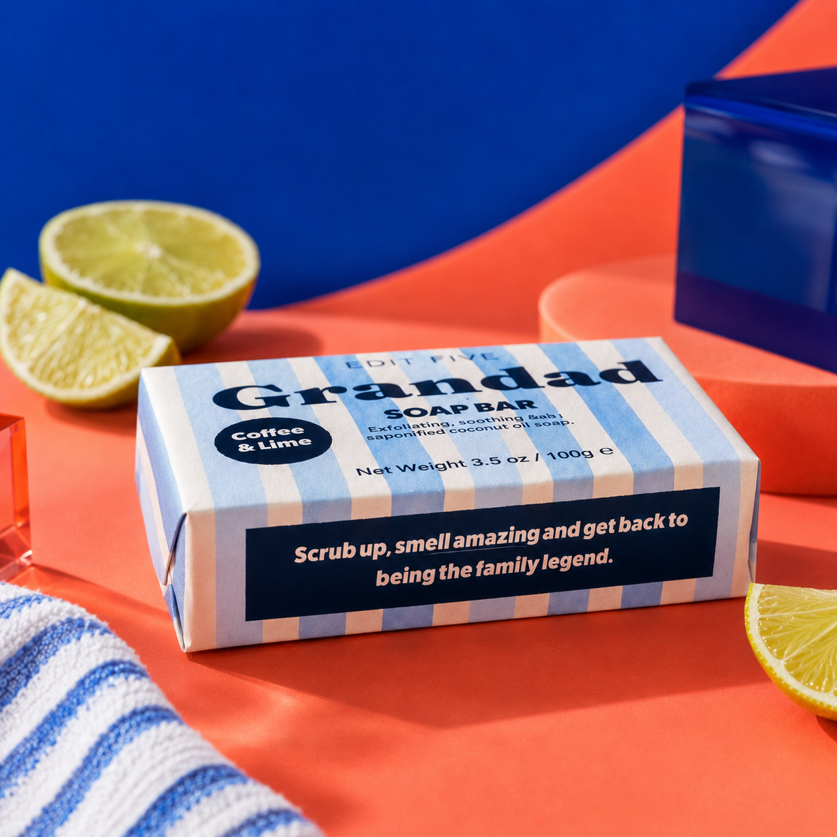 A boxed soap bar labeled 'Grandad' with 'Coffee & Lime' scent, shown on a bright orange surface with sliced lime and a blue-and-white striped towel nearby. The product box is the main subject, and no person or animal is present. Edit Five coffee and lime soap bar designed especially for Grandad, featuring natural essential oils and colourful packaging—a practical birthday gift, Christmas present or Father’s Day gift.