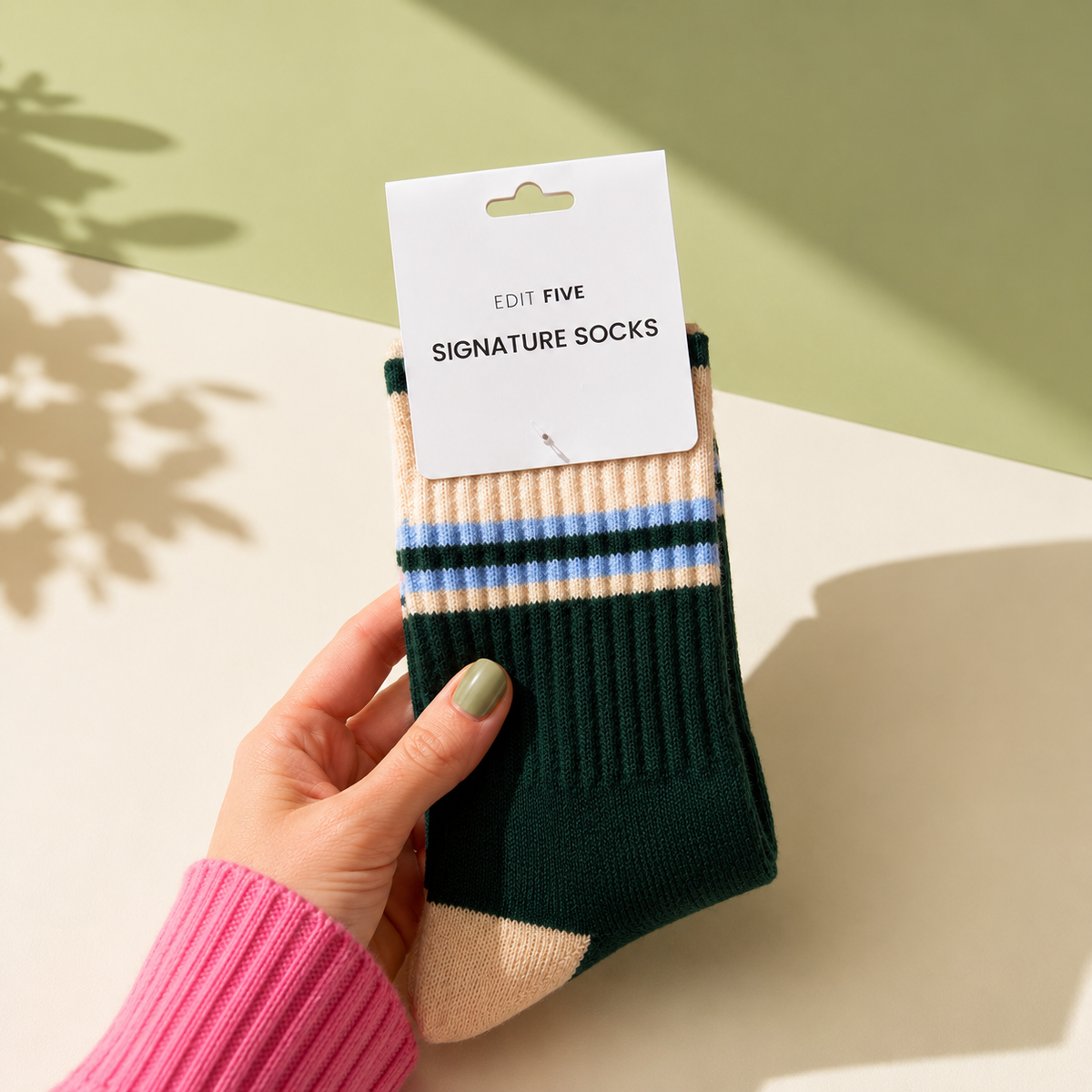 A hand holds a pair of dark green crew socks with beige and light blue stripes, still attached to a white paper hanger card that reads 'EDIT FIVE SIGNATURE SOCKS'. The background is a styled studio setup with a light beige surface and a sage green wall, featuring soft leaf shadows. Women’s green, peach and blue striped socks made from a premium soft-touch wool blend, with a warm, breathable and comfortable stretch fit for everyday wear.