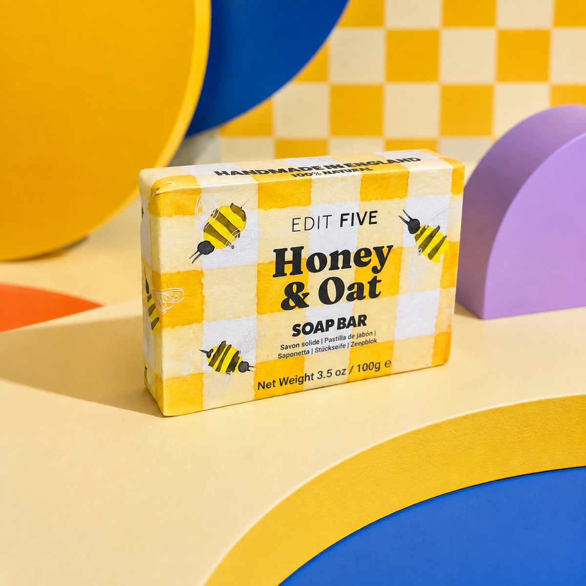 A boxed soap bar labeled 'EDIT FIVE Honey & Oat SOAP BAR' standing upright on a colorful geometric set with yellow, blue, purple, and beige shapes. Honey and oat soap bar, handmade in England using traditional methods, with a naturally comforting fragrance and rich, creamy lather for gentle everyday cleansing.