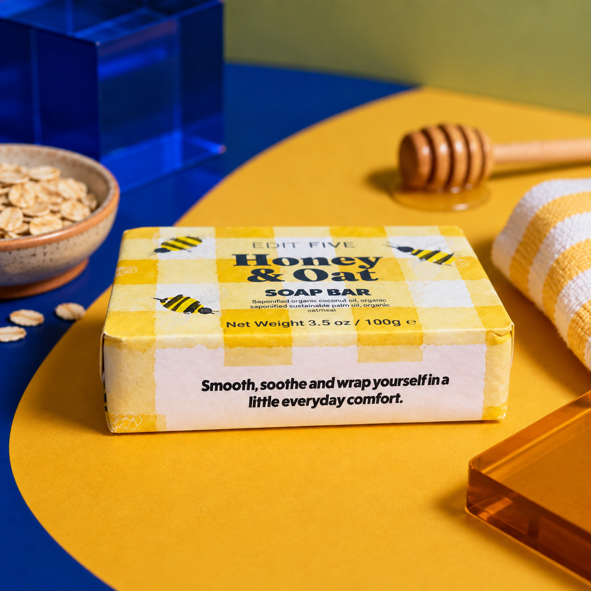 A bar of Edit Five Honey & Oat soap bar in a yellow and white gingham box, styled on a yellow and blue surface with honey-themed props including a wooden dipper, honeycomb, oats, and a folded yellow-and-white cloth. Edit Five honey and oat soap bar featuring nourishing honey and comforting oats, beautifully packaged as a thoughtful pampering gift, bathroom essential or gift-hamper addition.