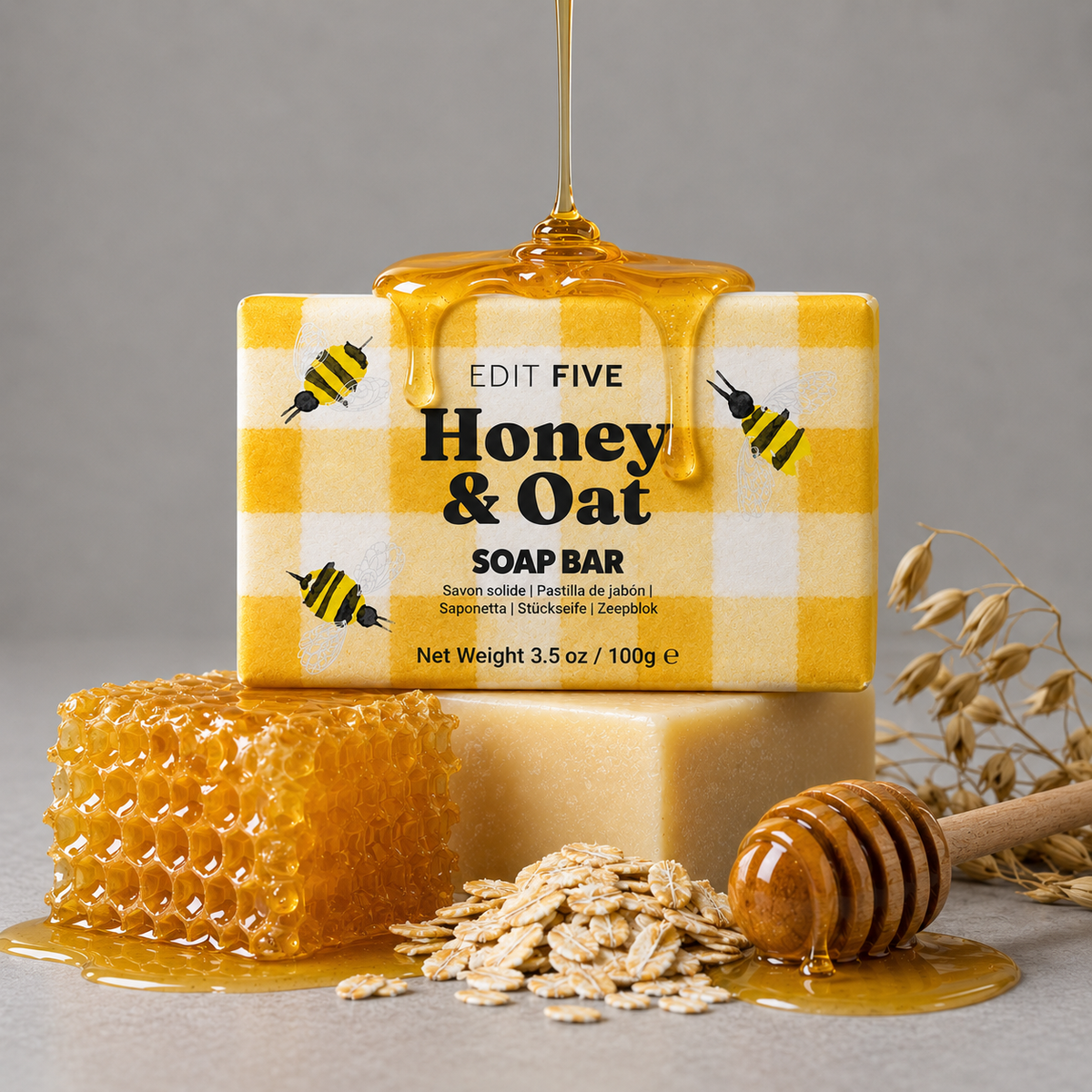 A boxed Honey & Oat soap bar sits on top of a plain soap bar, with honey dripping from above. The front box is yellow-and-white checkered with bumblebee illustrations and the text 'EDIT FIVE Honey & Oat SOAP BAR. Luxury handmade honey and oatmeal soap with a gentle cleansing formula, soft natural fragrance and creamy lather, perfect for anyone who enjoys comforting bath and body products.