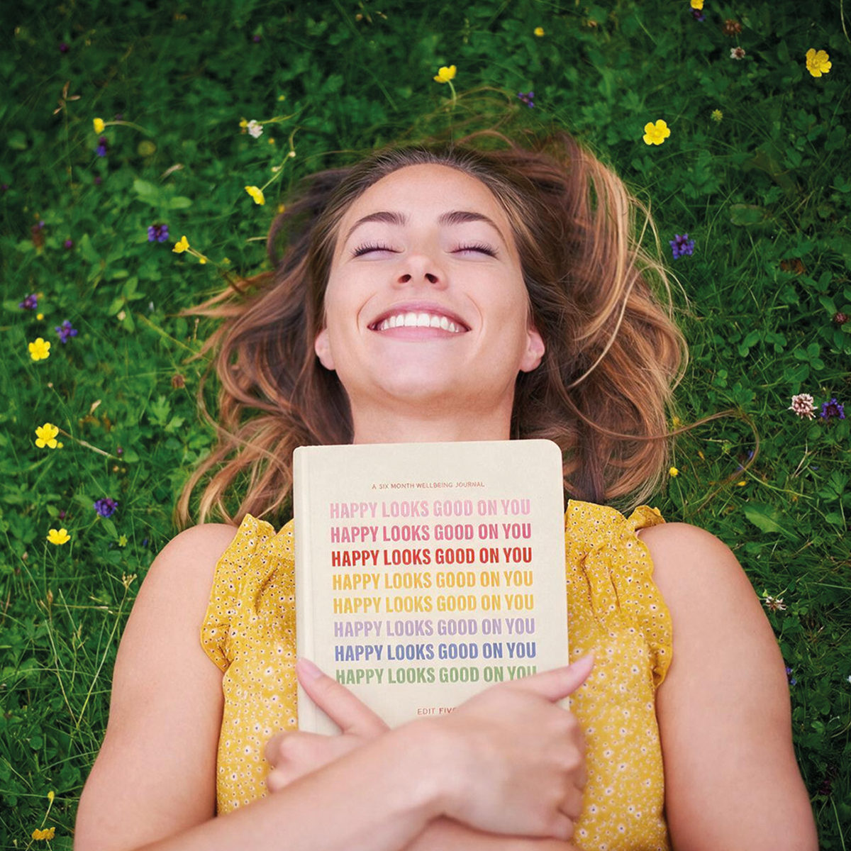 A woman lying on a grassy field holds up a book titled 'Happy Looks Good On You'. Mental health, 6 month wellbeing journal.