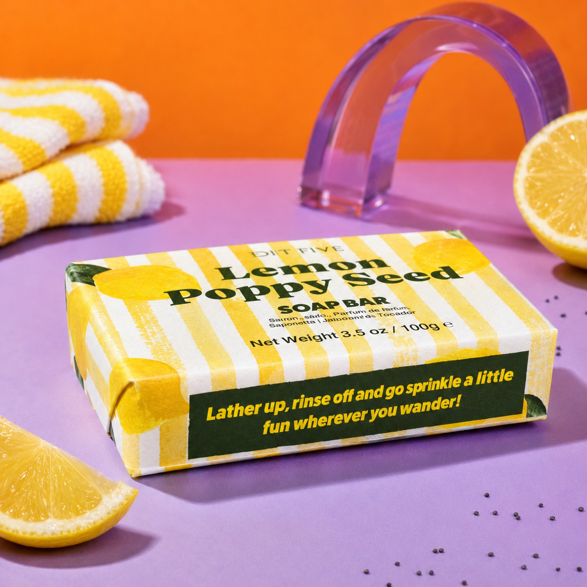 A boxed lemon poppy seed soap bar sits on a lavender surface with orange and yellow props. The package is yellow and white striped with green text reading 'Lemon Poppy Seed SOAP BAR' and a tagline on the front. Edit Five lemon and poppy seed exfoliating soap bar with natural essential oil, ideal for energising morning showers, daily bathing, self-care routines and citrus lovers.