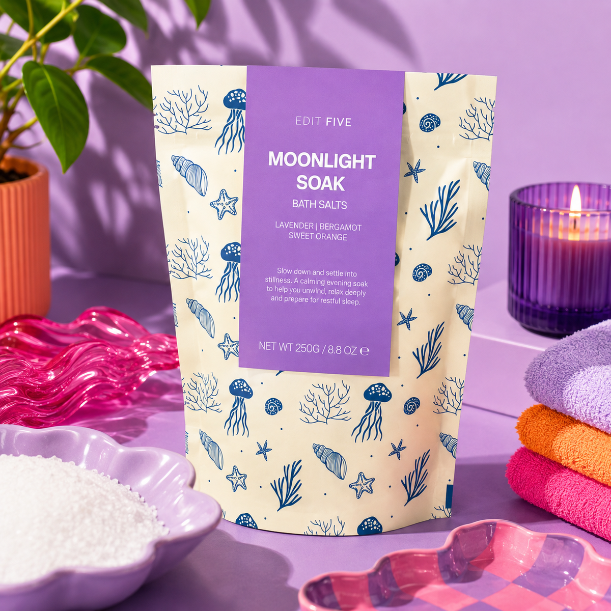 A stand-up pouch of Edit Five Moonlight Soak bath salts, featuring a lavender label and a cream background with blue jellyfish and starfish illustrations. The pouch is styled on a purple surface with a pink glass dish of white bath salts, folded pastel towels, and a purple candle in the background. Vegan-friendly lavender bath salts handmade in Staffordshire with mineral-rich Dead Sea salts, Epsom salts and uplifting bergamot and sweet orange essential oils.