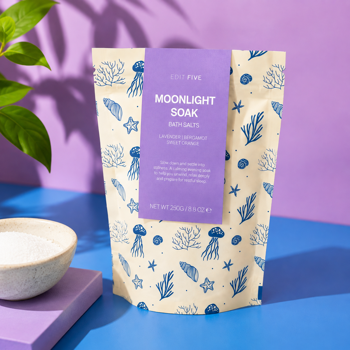 A stand-up pouch of Edit Five Moonlight Soak bath salts, featuring a lavender label and a cream background with blue sea-creature illustrations, styled on a blue and purple surface with a small bowl of white salts nearby. Moonlight Soak bath salts with Epsom and Dead Sea salts, scented with lavender, bergamot and sweet orange essential oils for calming evening baths and bedtime relaxation.