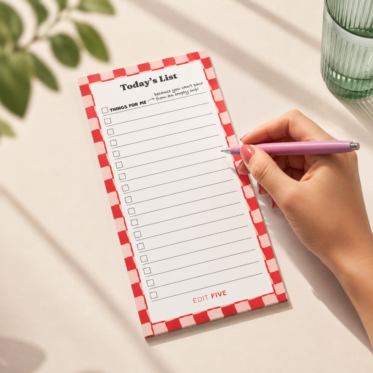 A hand with pink nails holds a lavender pen over a red-and-white checkered notepad labeled 'Today's List' on a light beige surface with soft shadows and a green glass in the background. Colourful “Today’s List” long daily planner notepad with lined sections for organising tasks, priorities and reminders. A bright, modern desk stationery essential for productivity, work, home organisation, study and everyday to-do lists.