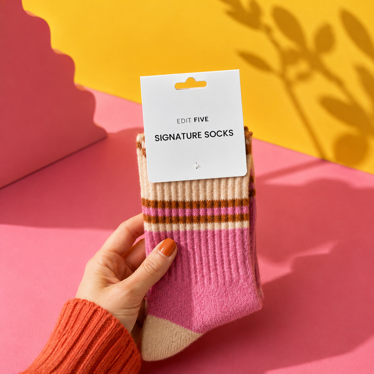 A hand wearing a rust-colored knit sweater holds a pair of pink and beige striped socks with a white paper tag that reads 'EDIT FIVE SIGNATURE SOCKS'. The background is a vibrant pink and yellow studio setup with leaf shadows. Women’s pink, peach and orange striped socks made from a soft, warm wool-blend fabric, with a comfortable stretch fit for colourful everyday wear.