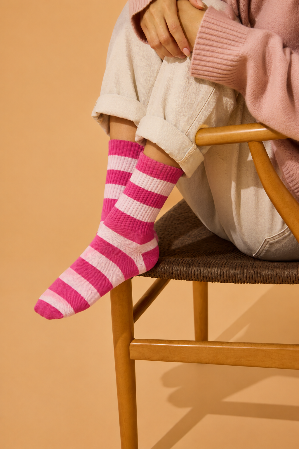 A person seated on a wooden chair wearing bright pink and white striped socks, off-white cuffed pants, and a light pink sweater against a warm beige background. Colourful women’s wool-blend socks featuring bold cherry pink and pale baby pink stripes—a fun fashion accessory, cosy winter essential and thoughtful gift for her.