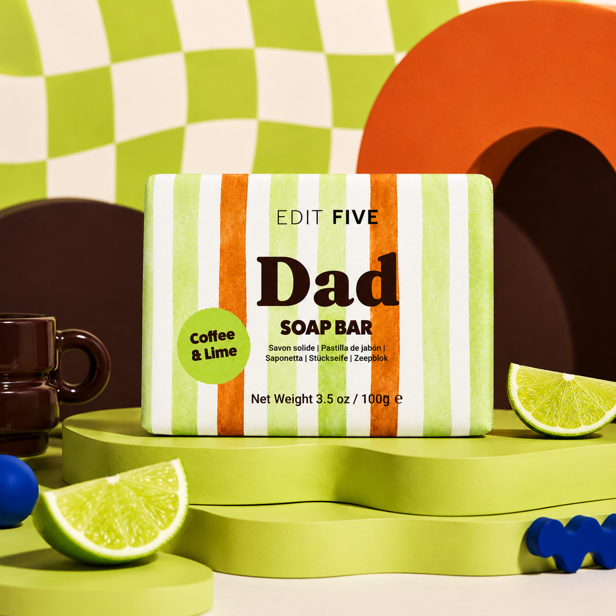 A boxed soap bar labeled 'EDIT FIVE Dad SOAP BAR' with a green, white, and orange striped package, styled on a bright green platform with lime wedges, a dark coffee mug, and abstract geometric props. Dad coffee and lime soap bar, handmade in England with natural lime essential oil, exfoliating coffee and bold gift-ready packaging—perfect for Father’s Day, birthdays or Christmas.